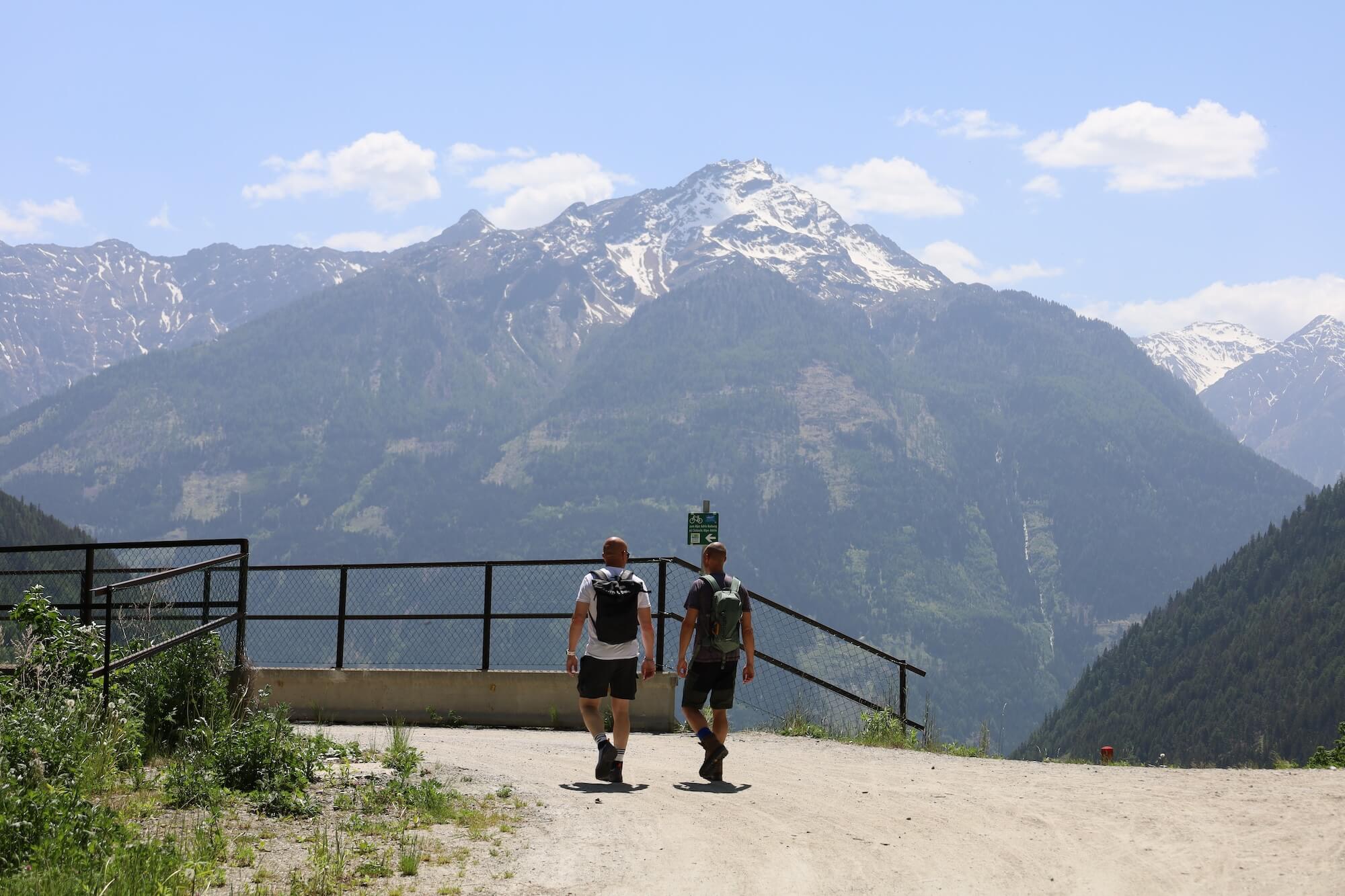 Twee wandelaars met rugzak aan metal railing op viewpoint overlooking massieve besneeuwde bergtop en diepe vallei